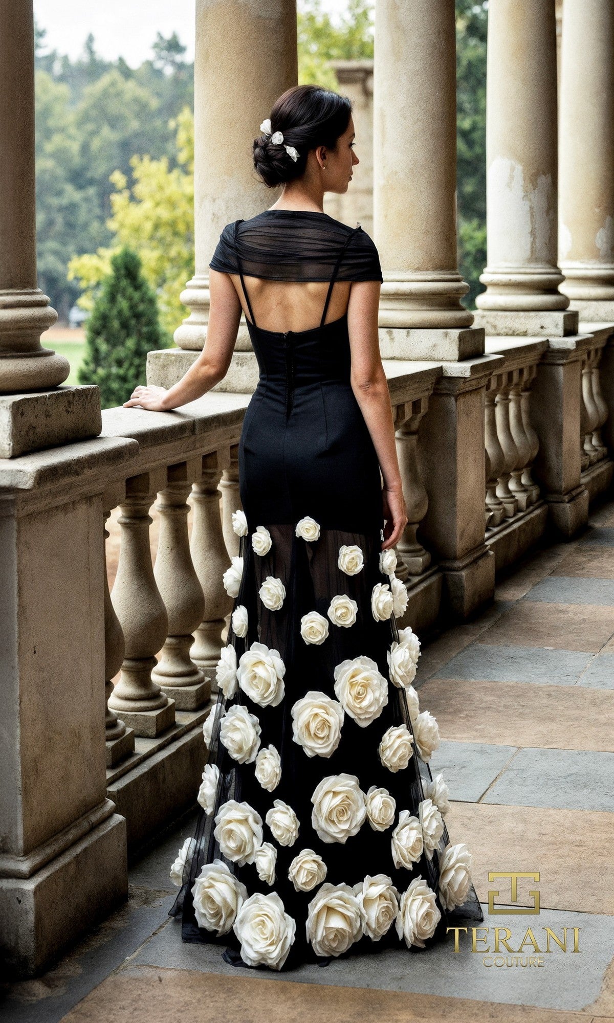 Wearing the Long Formal Dress: Terani 261P6098, featuring a sheer tulle skirt and oversized 3D floral appliques, a woman with an elegant updo stands on a stone terrace, facing away.